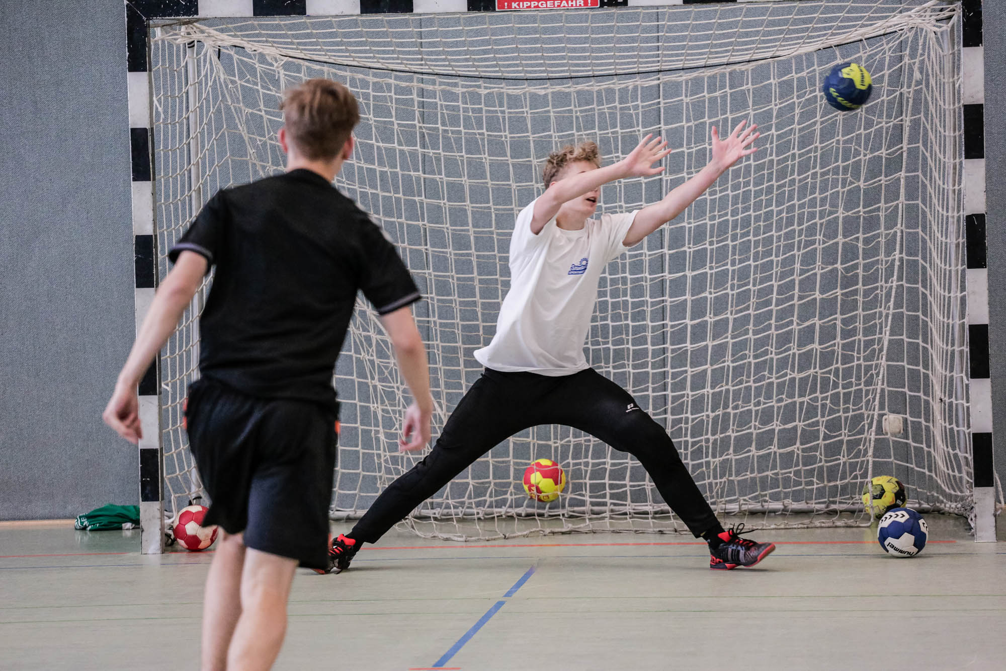 Das Bild zeigt eine Szene aus einem Handballspiel. Im Vordergrund sind zwei Jungen zu sehen. Einer der Jungen steht mit dem Rücken zur Kamera und trägt ein schwarzes T-Shirt und eine schwarze Hose. Er scheint gerade einen Wurf auszuführen. Der andere Junge steht im Tor und versucht, den Ball zu fangen. Er trägt ein weißes T-Shirt und eine schwarze Hose. Er hat die Arme weit geöffnet und versucht, den Ball abzuwehren. Der Ball ist gerade im Flug und nähert sich dem Tor. Im Hintergrund ist ein Handballtor zu sehen, das aus einem Netz und einem Rahmen besteht. Auf dem Boden liegen weitere Bälle. Die Halle hat graue Wände.