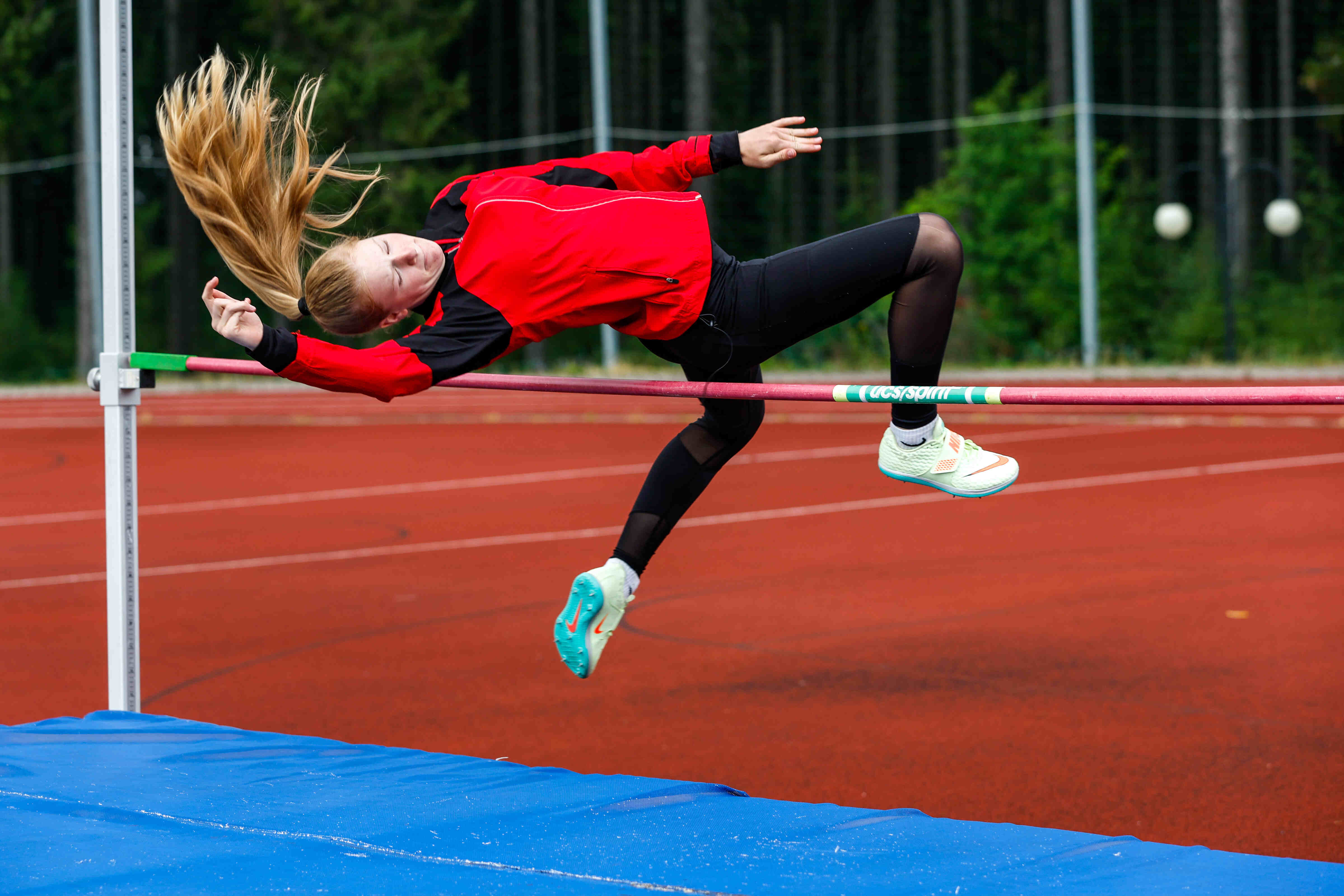 Das Bild zeigt ein Mädchen, das Hochsprung macht. Es ist in der Luft und versucht, über die Latte zu springen. Sie trägt eine rote Jacke, schwarze Leggings und Laufschuhe. Ihre Haare sind blond und fliegen in der Luft. Die Latte ist rot und grün. Das Bild wird auf einer roten Sportbahn vor einem grünen Hintergrund aufgenommen. Unter der Latte befindet sich eine blaue Matte.