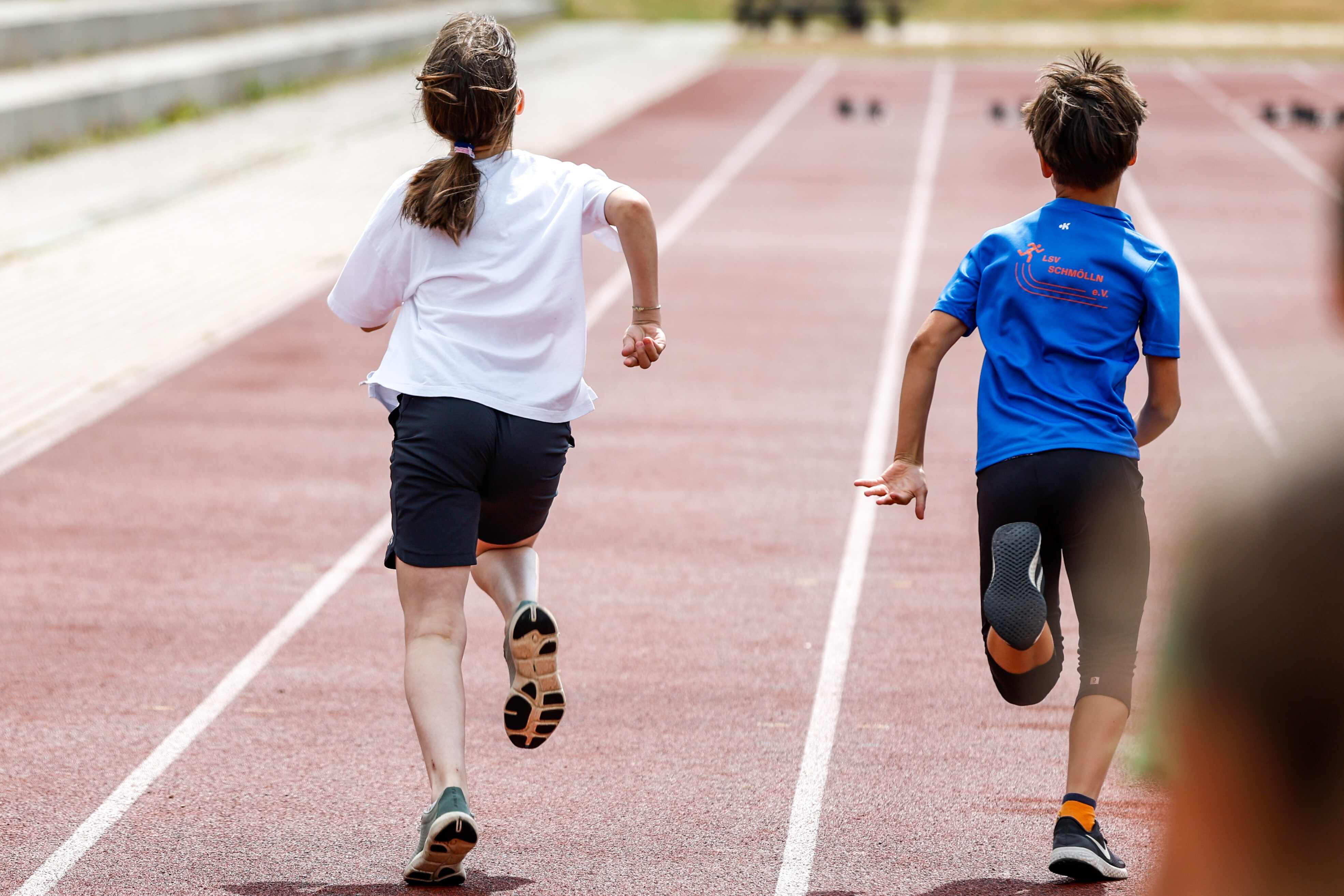 Das Bild zeigt zwei Kinder, die auf einer roten Laufbahn rennen. Beide sind von hinten zu sehen. Das Mädchen, das links auf dem Bild rennt, trägt ein weißes T-Shirt und schwarze Shorts. Sie hat ihr Haar zu einem Pferdeschwanz zusammengebunden. Ihre Arme sind angewinkelt. Der Junge, der rechts auf dem Bild rennt, trägt ein blaues T-Shirt mit einem Logo auf der Rückseite. Er trägt schwarze Shorts und schwarze Schuhe mit orangefarbenen Socken. Seine Arme sind angewinkelt. Die Laufbahn hat weiße Markierungen.