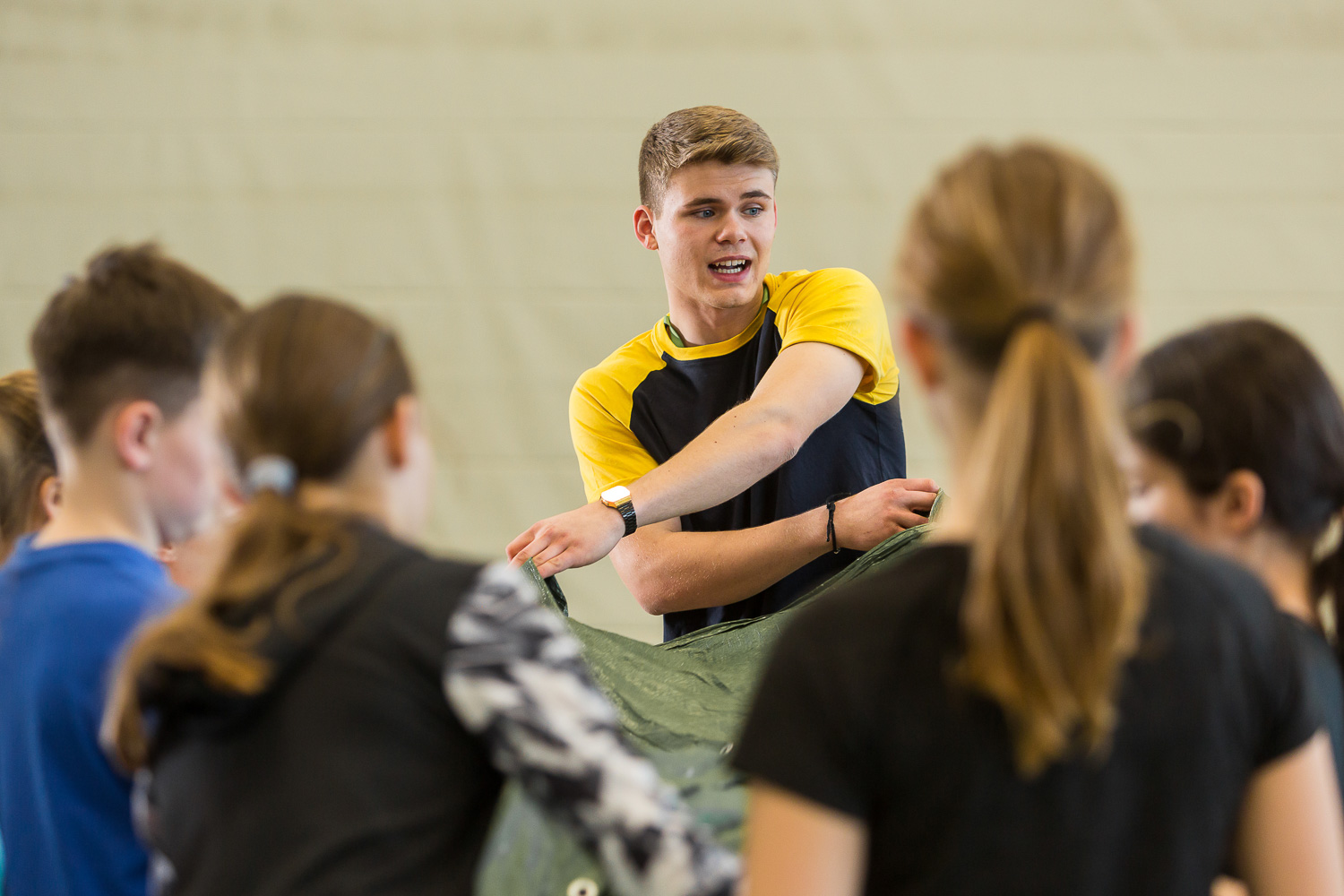 Das Bild zeigt eine Gruppe von Menschen, die in einem hellen Raum zusammenkommen. Im Mittelpunkt steht ein junger Mann, der sich anscheinend an eine Gruppe von Kindern wendet, die ihm gegenüberstehen. Er trägt ein gelb-schwarzes T-Shirt und eine Armbanduhr. Er hält eine grüne Plane in seinen Händen. Die Kinder um ihn herum sind nicht im Detail zu erkennen, aber es scheint, dass sie ihm zuhören.
