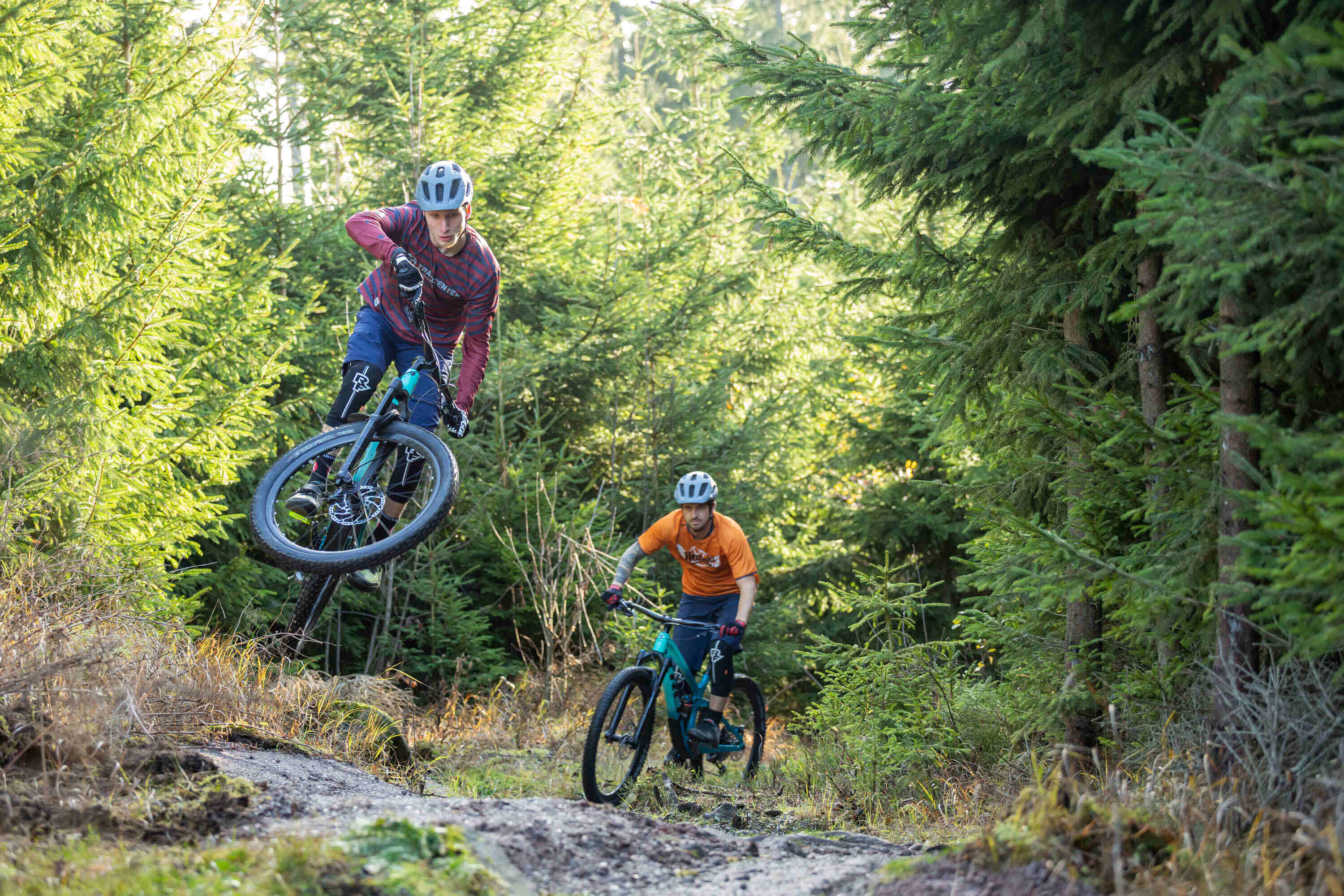 Sportpark Rabenberg: TrailCenter® Rabenberg Das Bild zeigt zwei Mountainbiker, die einen Waldweg hinunterfahren. Der erste Biker, der sich im Vordergrund befindet, ist in der Luft und springt über einen Hügel. Er trägt einen Helm, eine rot-schwarz gestreifte Jacke, blaue Shorts und Knieschützer. Sein Fahrrad ist schwarz und türkis. Der zweite Biker befindet sich hinter dem ersten Biker auf dem Weg. Er trägt einen Helm, ein orangefarbenes T-Shirt und schwarze Shorts. Sein Fahrrad ist ebenfalls schwarz und türkis. Der Hintergrund des Bildes ist ein Wald mit grünen Bäumen. Der Himmel ist hell, und das Sonnenlicht scheint durch die Bäume. Das Bild ist gut beleuchtet und die Farben sind lebendig. Es vermittelt ein Gefühl von Bewegung und Abenteuer.