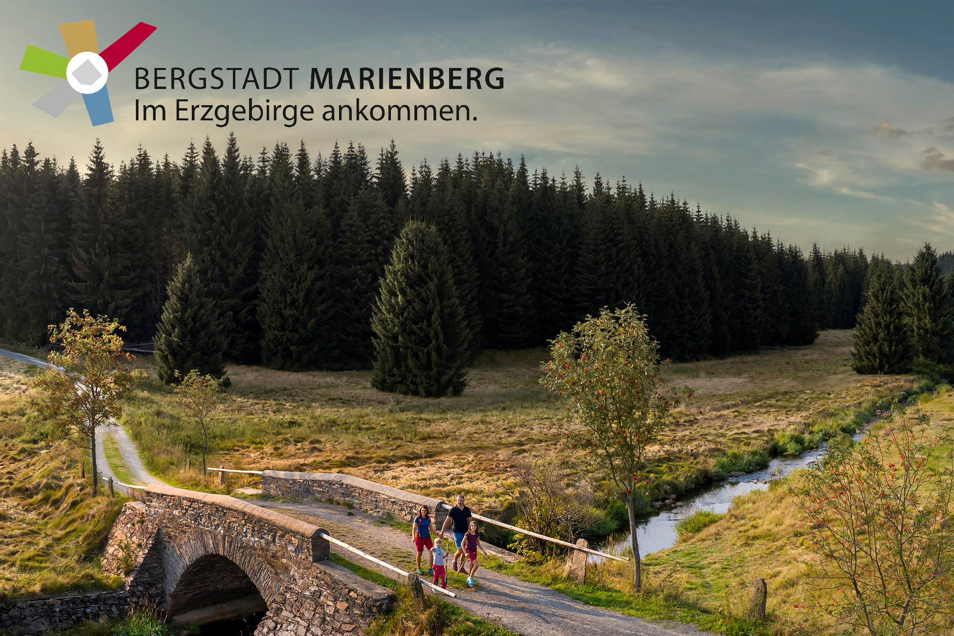 Sportpark Rabenberg EURORANDO 2026 Aussteller: Stadt Marienberg; © Uwe Meinhold Familie überquert Steinbrücke im Erzgebirge neben einem Bach und einem Wald; Logo Stadt Marienberg in oberer linker Ecke
