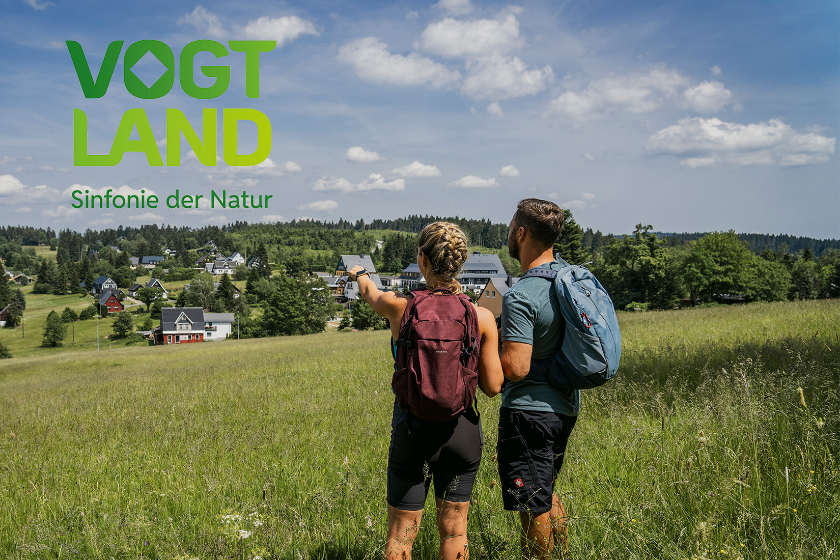 Sportpark Rabenberg EURORANDO 2026 Aussteller: Tourismusverband Vogtland; © Dirk Rückschloß | pixore photography Ein Paar mit Rucksäcken blickt auf ein Dorf und Bergpanorama. Linke obere Ecke Logo "Vogtland Sinfonie der Natur"
