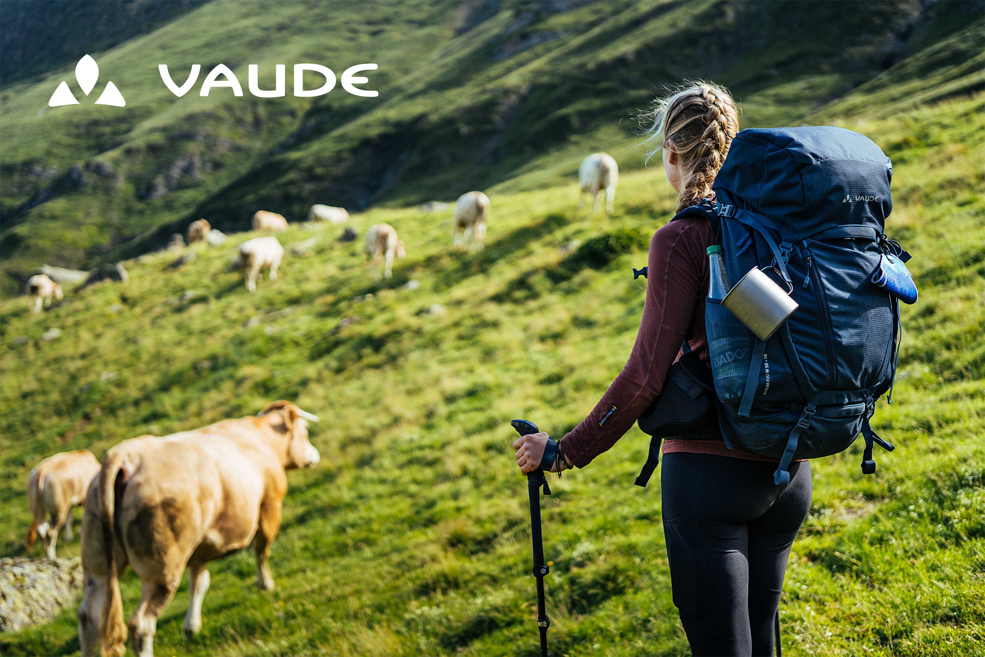 Sportpark Rabenberg EURORANDO 2026 Aussteller: VAUDE Wanderin mit Rucksack und Wanderstock geht über eine Bergwiese mit Kühen und Schafen. Vaude Logo oben links.