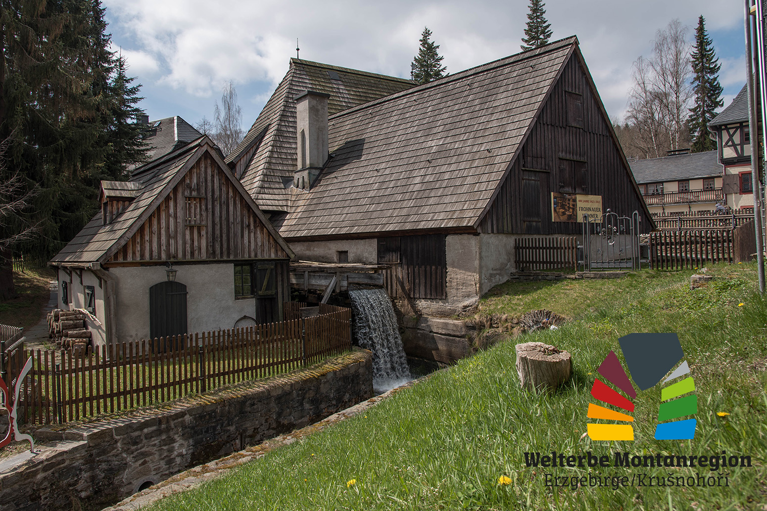 Sportpark_Rabenberg EURORANDO 2026 Aussteller: Welterbe Montanregion Erzgebirge; Historisches Gebäude mit Wasserrad, Wasserfall und Schindeldach, umgeben von Bäumen und Grünflächen.