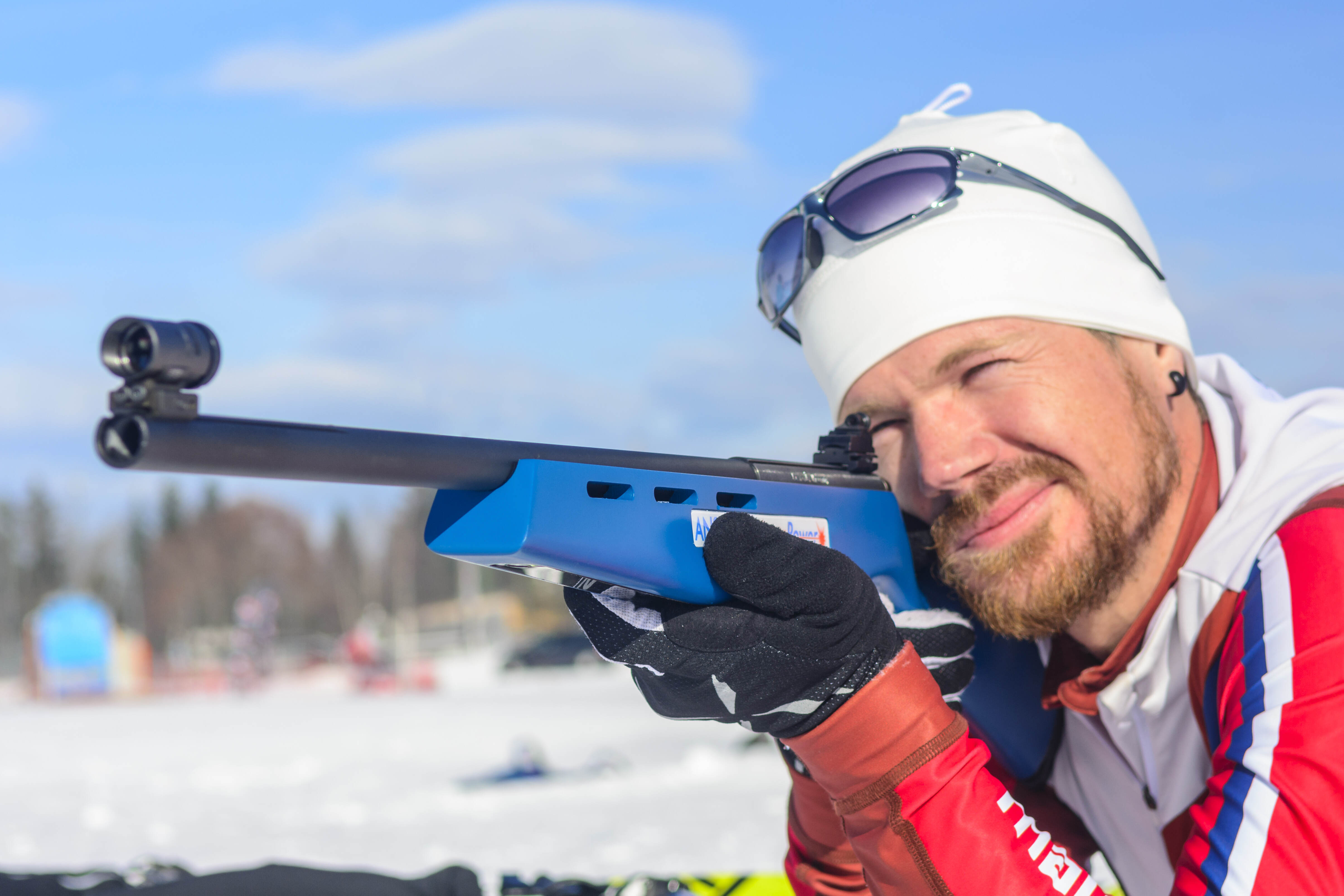 Das Bild zeigt eine Person, die mit einem Gewehr zielt. Die Person trägt eine weiße Mütze, Sonnenbrille, eine rot-weiß-blaue Jacke und schwarze Handschuhe. Das Gewehr ist blau und schwarz. Im Hintergrund ist Schnee und ein blauer Himmel zu sehen.