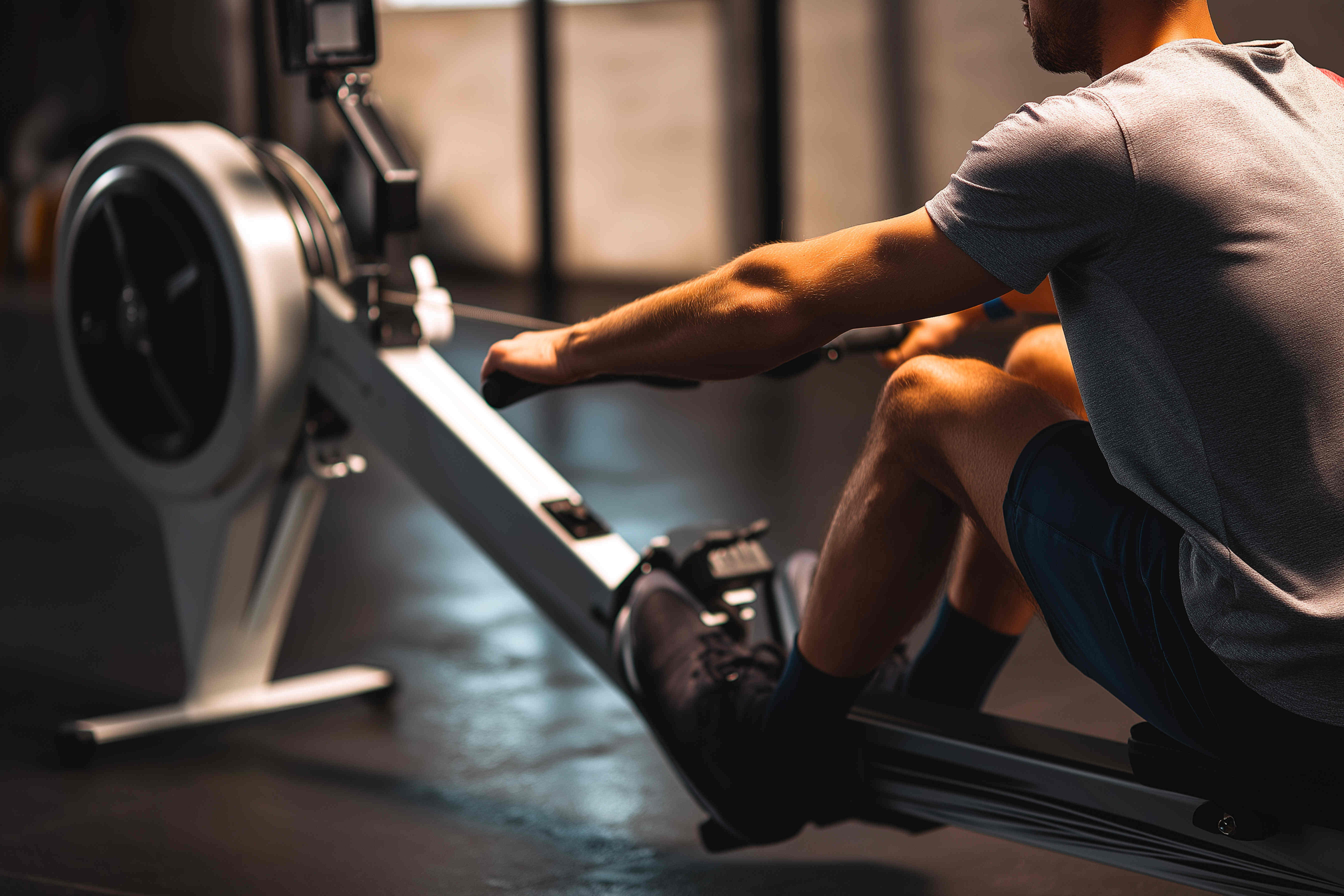 Ein Mann trainiert auf einem Rudergerät. Nur sein Oberkörper und seine Beine sind im Bild zu sehen. Er trägt ein graues T-Shirt und blaue Shorts. Seine Hand hält den Griff des Rudergeräts. Seine Beine sind in den Fußstützen des Geräts fixiert. Das Rudergerät ist weiß und schwarz.