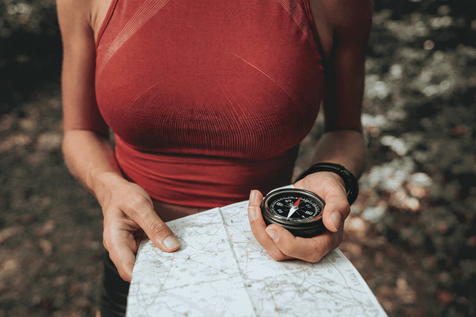 Das Bild zeigt eine Person, die einen Kompass und eine Karte in den Händen hält. Die Person trägt ein rotes Tanktop. Der Hintergrund ist unscharf und zeigt eine grüne, waldähnliche Umgebung.
